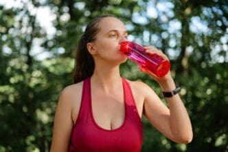 A woman in a red sports outfit drinks from a pink water bottle in a forest.
