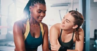 Two female friends chat and smile while discussing a workout plan in a gym.