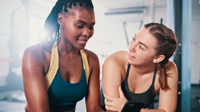 Two female friends chat and smile while discussing a workout plan in a gym.