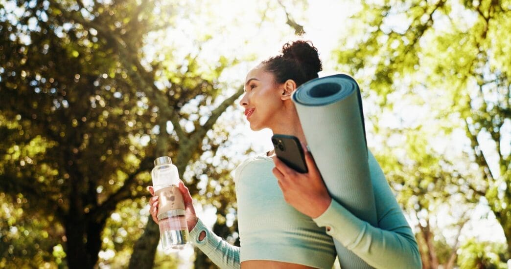 Woman smiling with a yoga mat, water bottle, and phone in a park, ready for a workout.