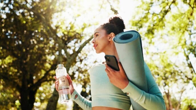 Woman smiling with a yoga mat, water bottle, and phone in a park, ready for a workout.