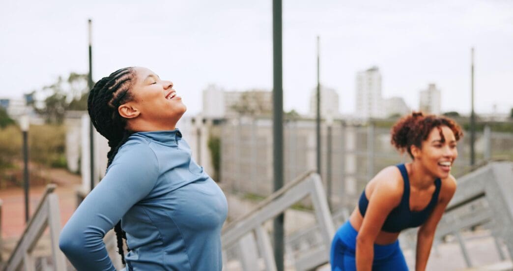 A group of tired, smiling women take a break together after a workout in a city setting.