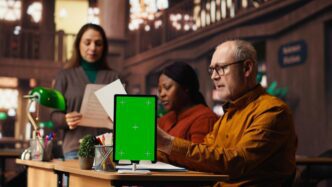 Diverse group of people, some using laptops with a green screen, study together in a public library.