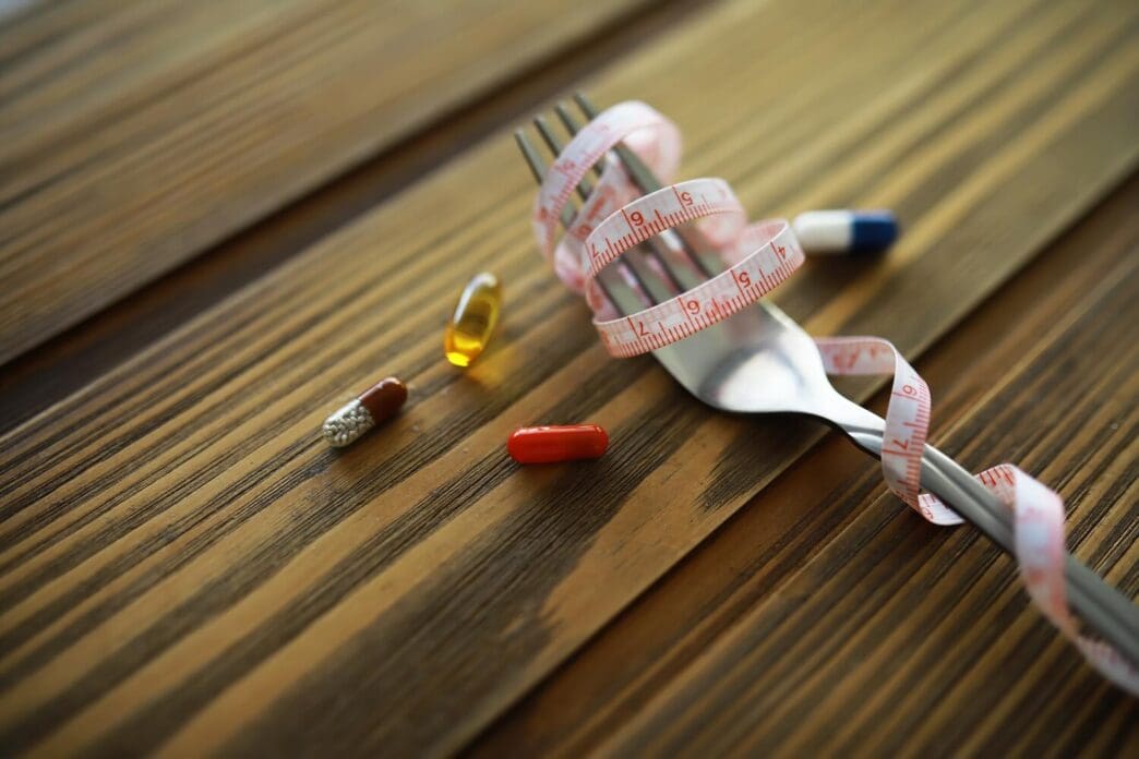 Fork with measuring tape wrapped around it, and pills arranged on a wooden table, representing diet and health.