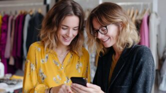 Two friends shop together in a boutique, sharing style ideas on their mobile phones.