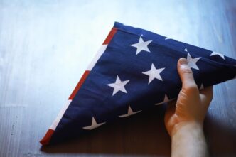 A person's hand gently holds a folded American flag on a rustic wooden surface, likely for a Memorial Day or Veterans Day observance.