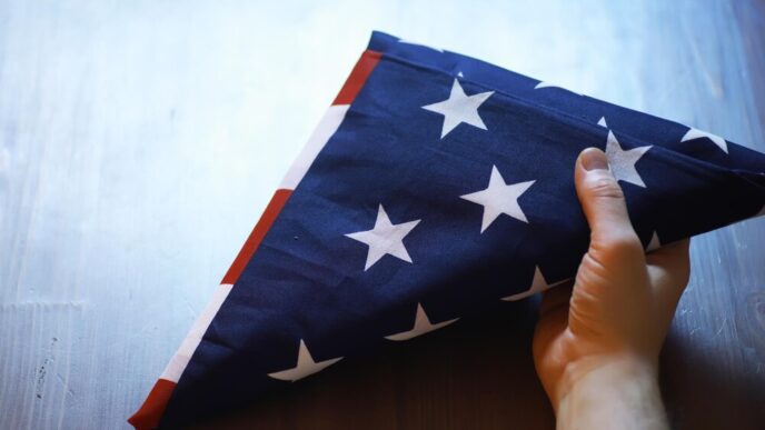 A person's hand gently holds a folded American flag on a rustic wooden surface, likely for a Memorial Day or Veterans Day observance.
