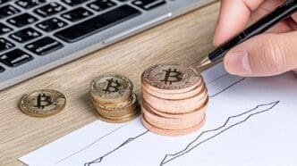 A hand holds a pen near Bitcoin coins and a financial chart on a table.