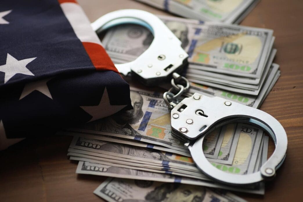 Handcuffs rest on top of a pile of dollar bills, with an American flag in the background, representing legal and financial themes.
