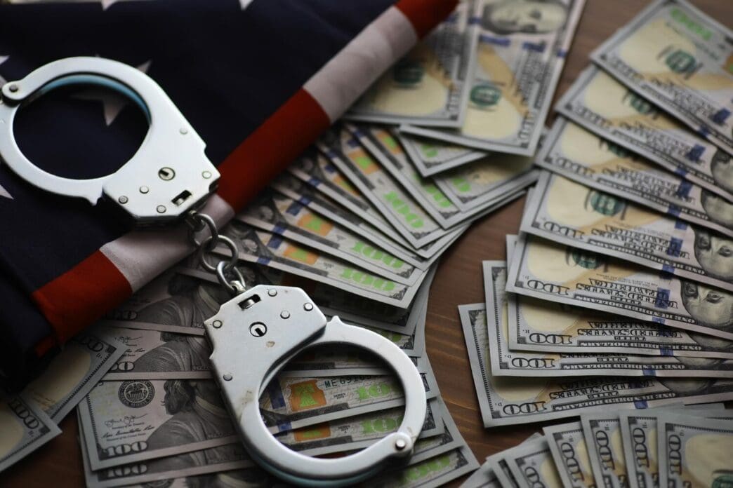 Handcuffs rest on a surface next to a US flag and piles of dollar bills, symbolizing financial crime and law enforcement.