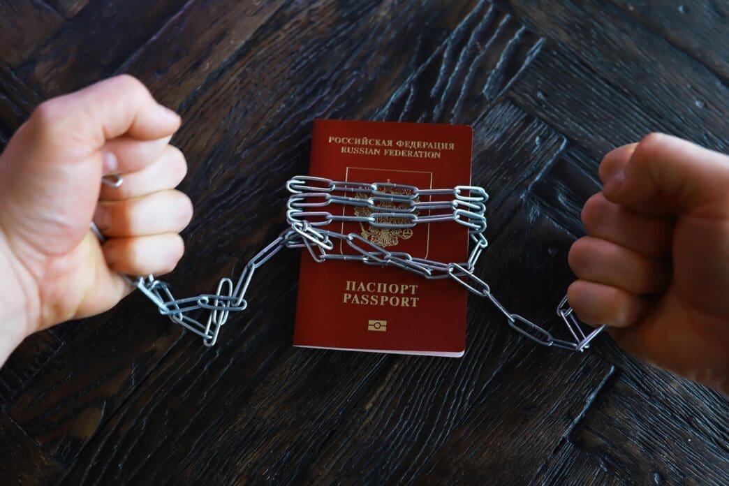 Hands in chains rest on a wooden table next to a Russian passport, symbolizing restriction and lack of freedom.