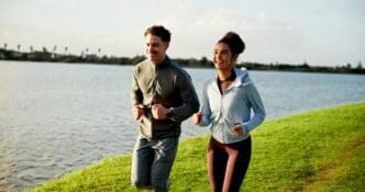 A smiling man and woman run together on a grassy field near a lake, likely for a workout or marathon training.