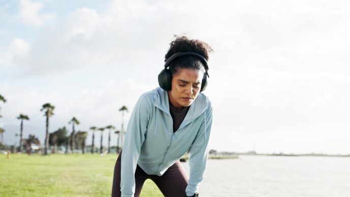 A woman wearing headphones rests on the edge of a lake, appearing exhausted after a workout.