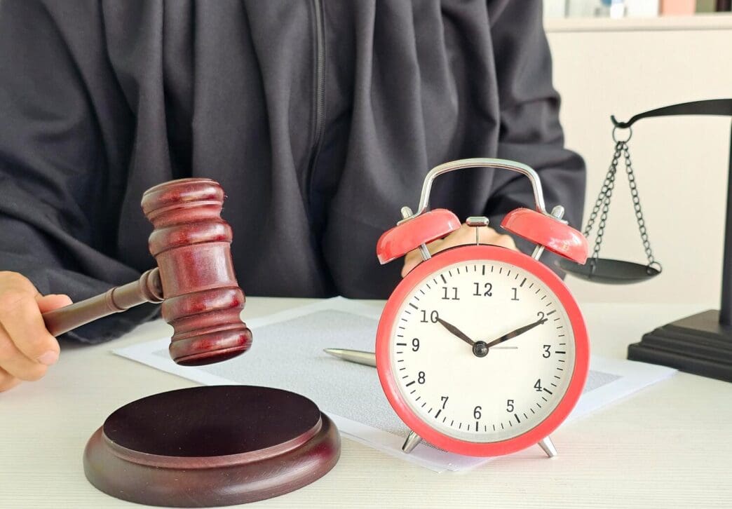 A judge sits at a courtroom table with a gavel, an alarm clock, and a scale of justice.