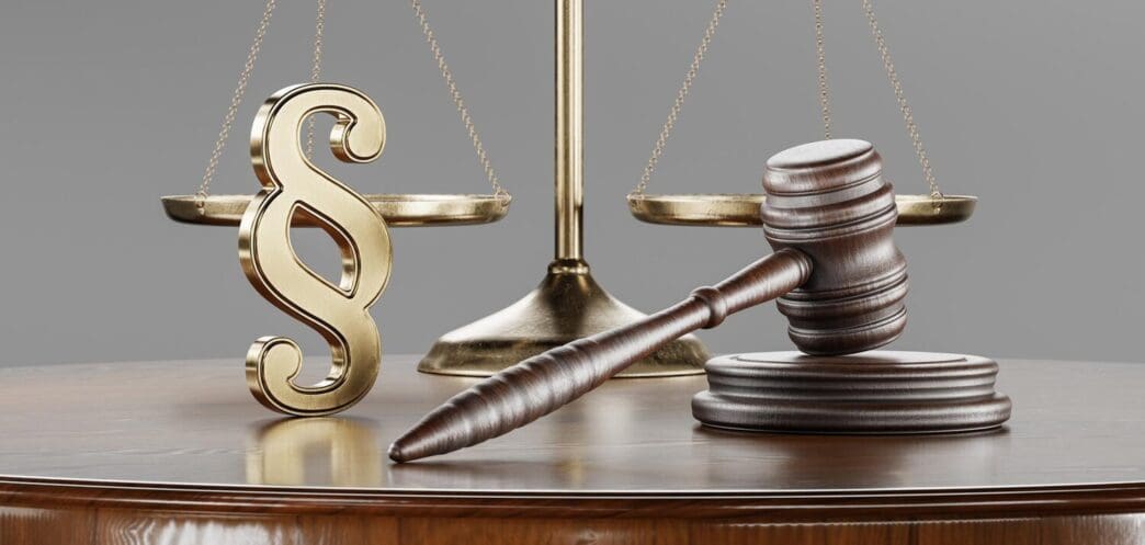 A gavel rests on a table beside a set of scales, a book, and a paragraph symbol, representing legal concepts.
