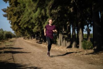 A man in a maroon athletic shirt runs along a dirt trail through a pine forest.