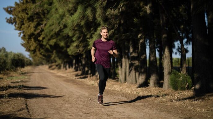 A man in a maroon athletic shirt runs along a dirt trail through a pine forest.