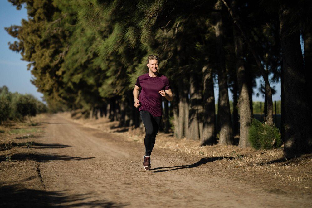 A man in a maroon athletic shirt runs along a dirt trail through a pine forest.