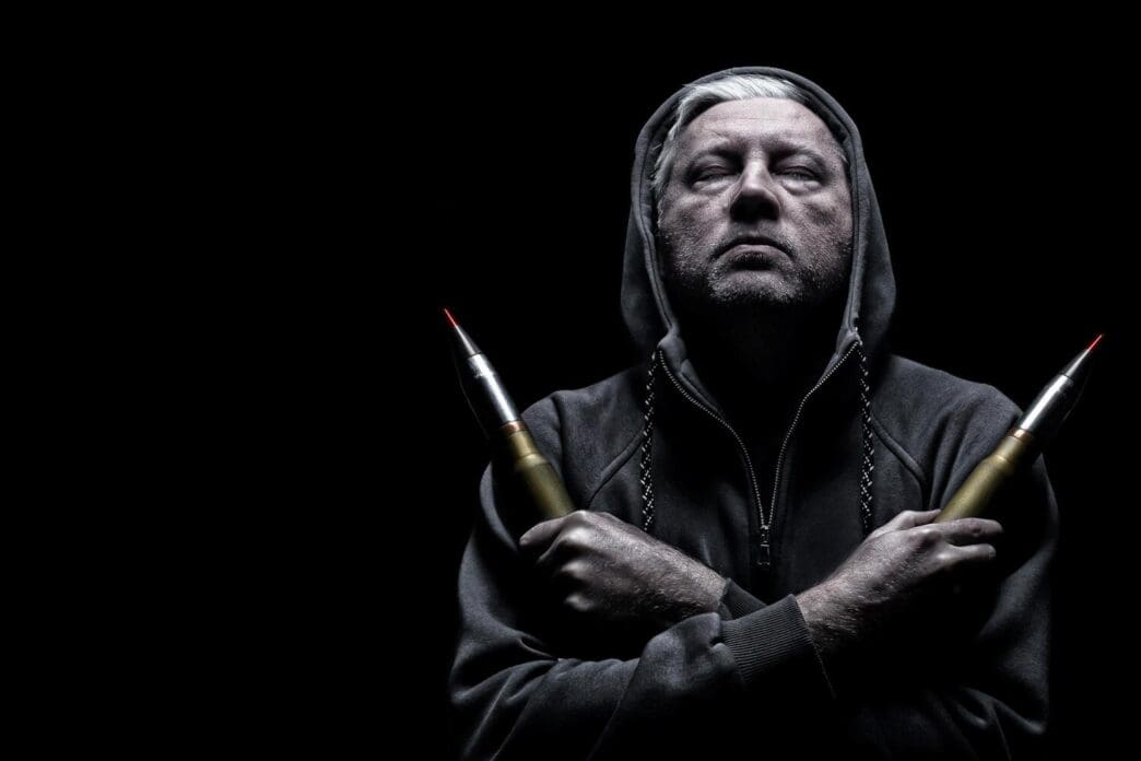 A man in a hoodie poses with bullets in dramatic lighting, taking a selfie.