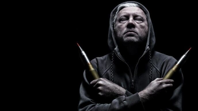 A man in a hoodie poses with bullets in dramatic lighting, taking a selfie.