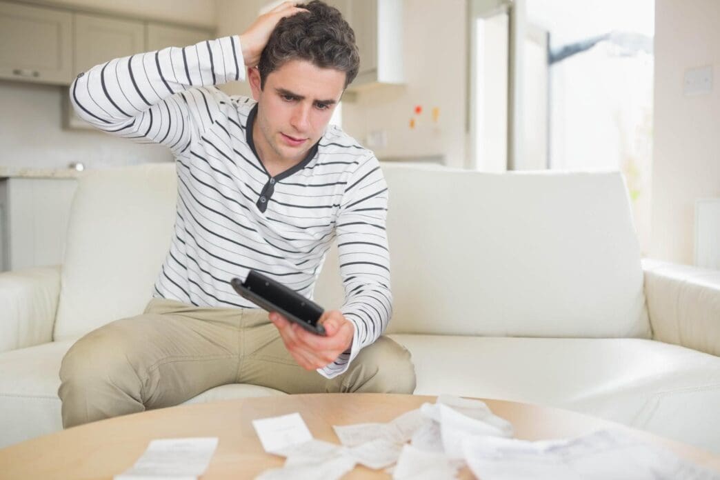Man on a sofa at home uses a calculator while sorting through receipts on a coffee table.