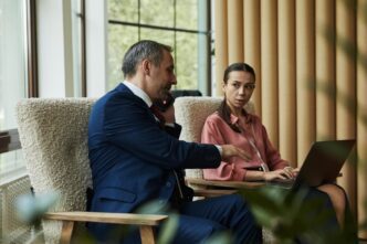 A middle-aged, light-skinned man speaks on the phone while standing near a woman using a laptop.