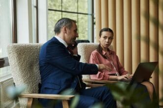 A middle-aged, light-skinned man speaks on the phone while standing near a woman using a laptop.
