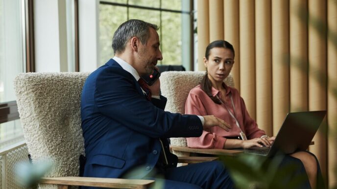 A middle-aged, light-skinned man speaks on the phone while standing near a woman using a laptop.