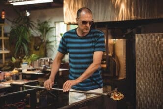 A middle-aged man stands at a glassblowing bench, gripping a blowpipe with molten glass near a furnace.