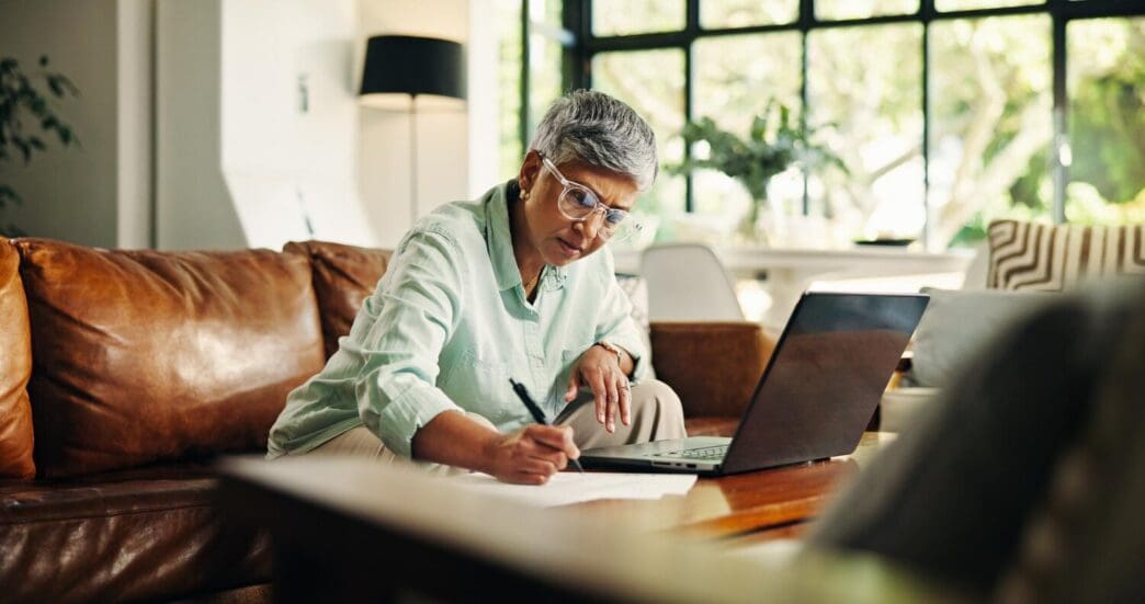 An elderly woman reviews financial documents on a laptop in her living room, possibly for debt management, budgeting, or insurance applications.