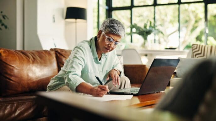 An elderly woman reviews financial documents on a laptop in her living room, possibly for debt management, budgeting, or insurance applications.