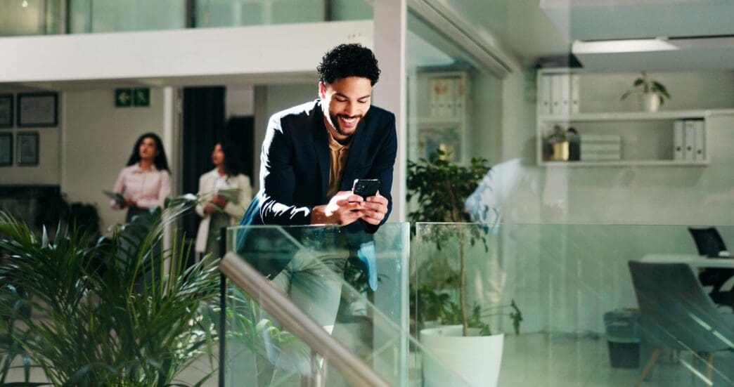 A businessperson in an office smiles while reading an email on a phone, possibly reviewing a sustainable project proposal.