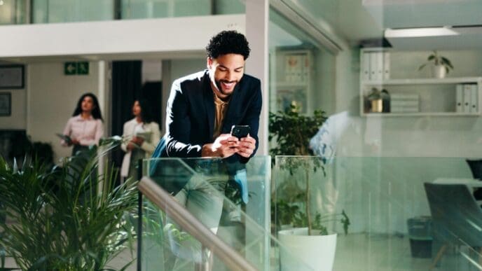 A businessperson in an office smiles while reading an email on a phone, possibly reviewing a sustainable project proposal.