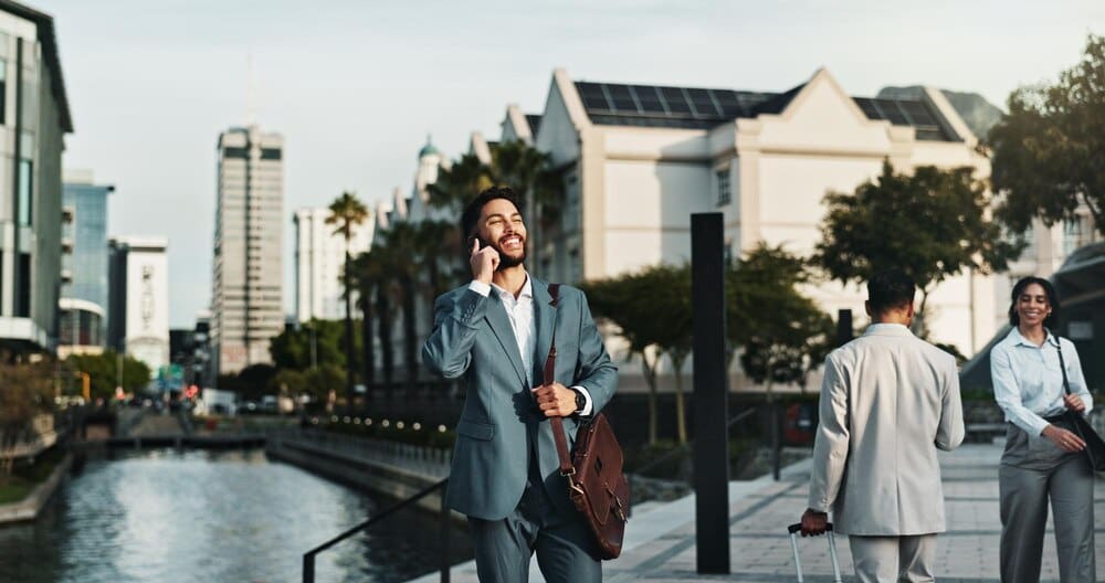 A professional man smiles while talking on his phone in a city setting, likely during a morning commute.