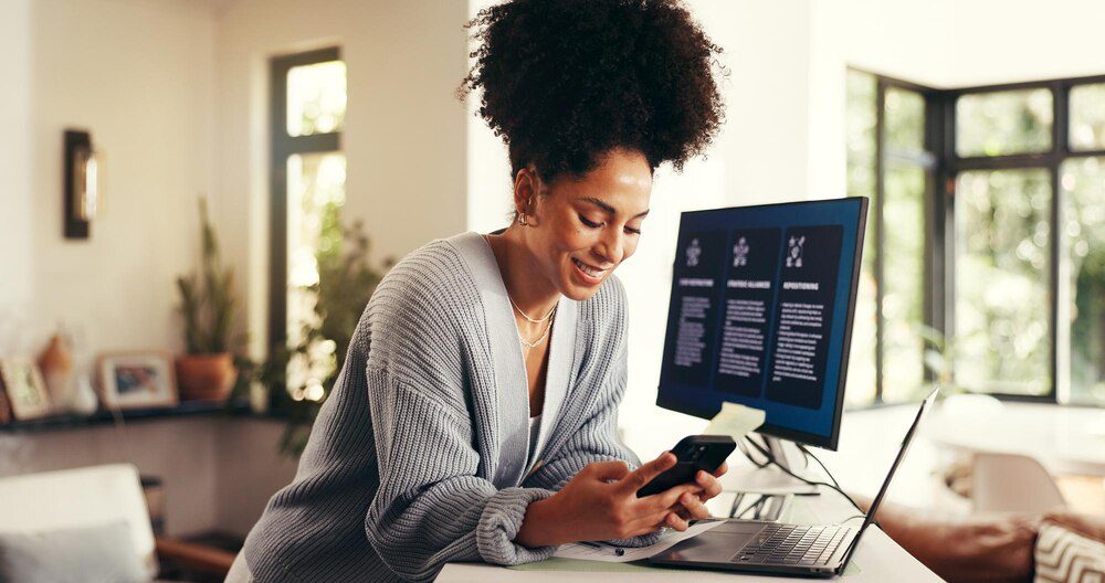 A woman works remotely on a phone and computer in a home office, likely for a consulting business, startup, or project management.