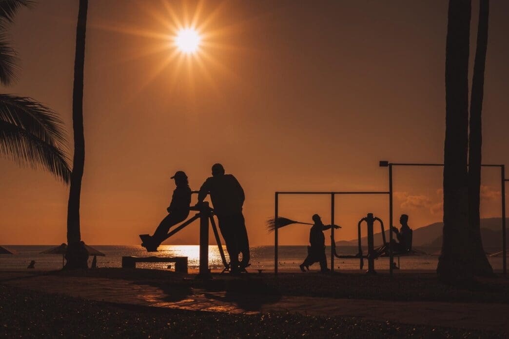 Silhouettes of elderly Vietnamese people exercise on horizontal bars at a sports ground by the sea at sunrise.