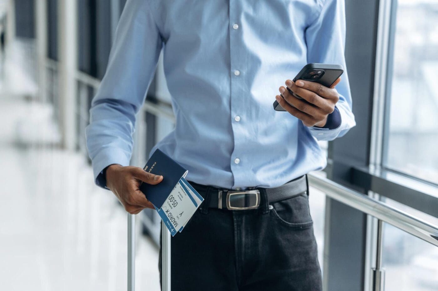A male tourist in an airport corridor holds a smartphone and tickets.