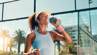 Woman on a tennis court takes a break to drink water from a sports bottle after a workout.
