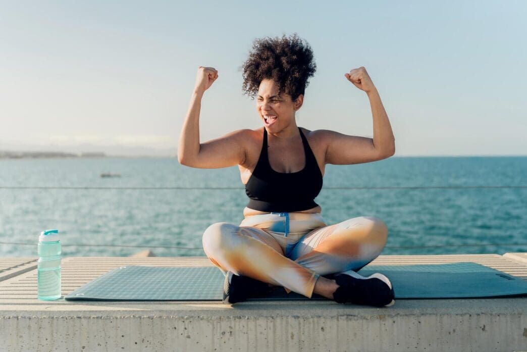 A muscular Hispanic woman flexes her arms, showcasing her strength after an outdoor workout by the sea.
