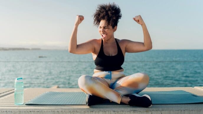 A muscular Hispanic woman flexes her arms, showcasing her strength after an outdoor workout by the sea.