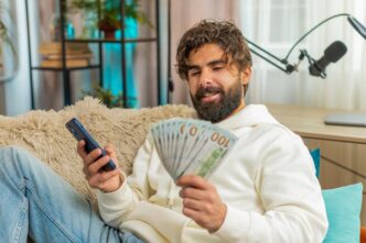 A young Indian man, holding a dollar bill and a smartphone, sits on a sofa and appears successful.