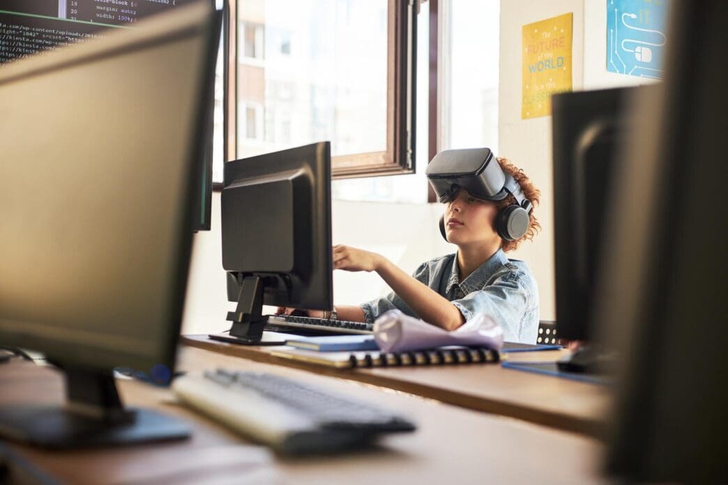 Teenage boy with fair skin wearing a VR headset is seated and coding on a computer.