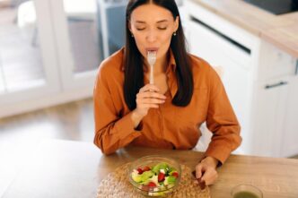 Woman eating a fresh vegetable salad and licking her fork while sitting at a table.