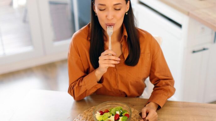 Woman eating a fresh vegetable salad and licking her fork while sitting at a table.