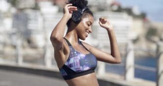 A Black woman smiles while dancing and wearing headphones in a city setting, likely listening to music or a podcast during a workout.