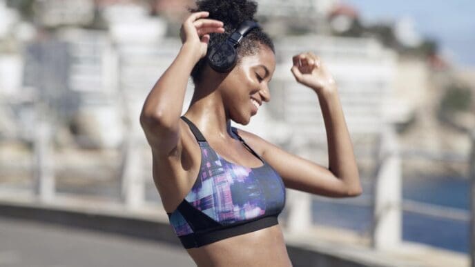 A Black woman smiles while dancing and wearing headphones in a city setting, likely listening to music or a podcast during a workout.