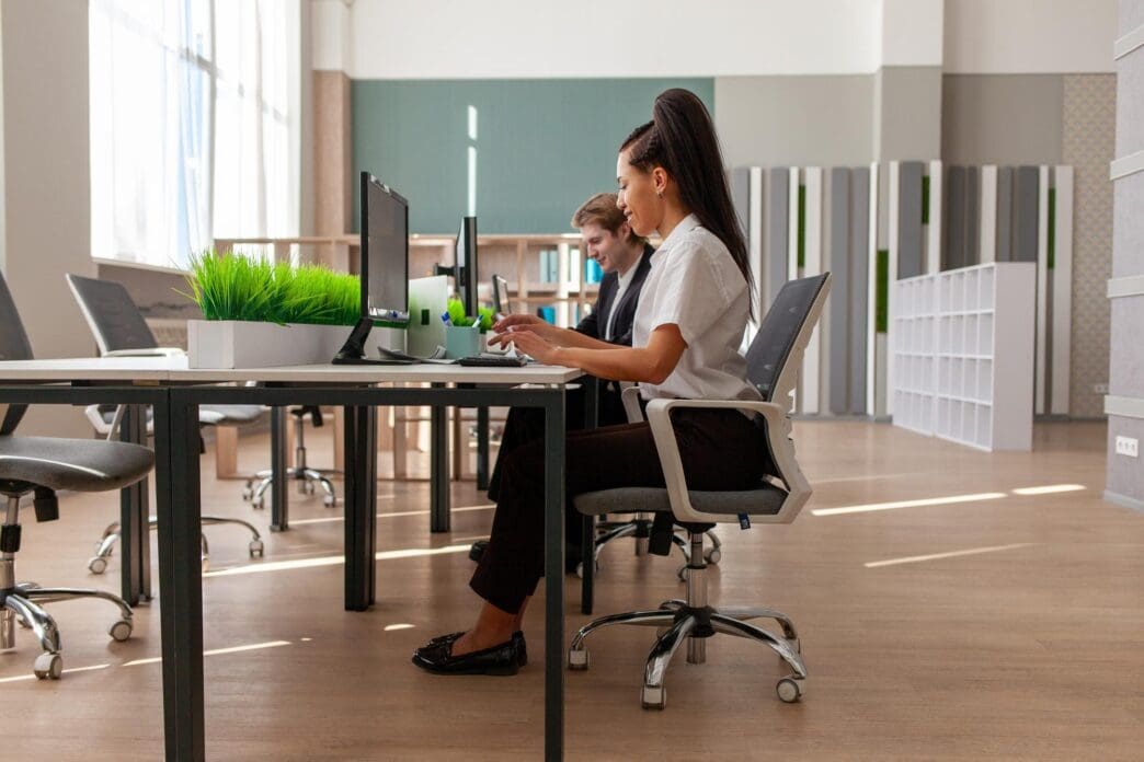 A woman and a young man in business suits sit at a desk in an office, typing on a computer.