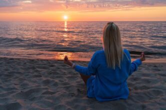 A woman in the lotus position meditates on a beach at sunset.