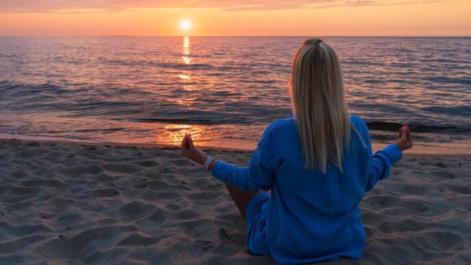 A woman in the lotus position meditates on a beach at sunset.