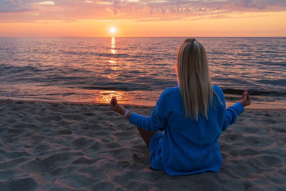 A woman in the lotus position meditates on a beach at sunset.